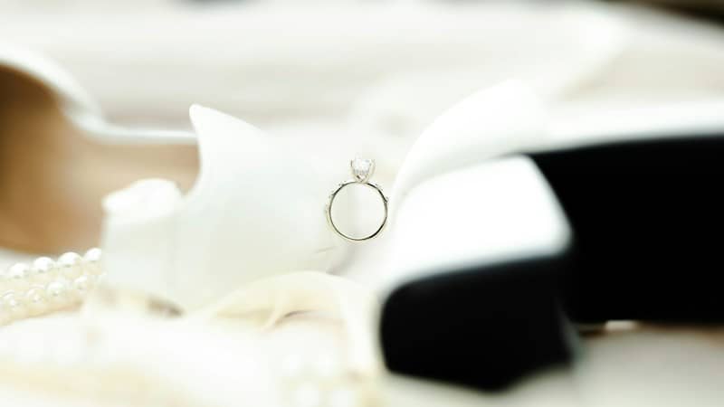 A diamond engagement ring resting on a white satin shoe with pearl accents