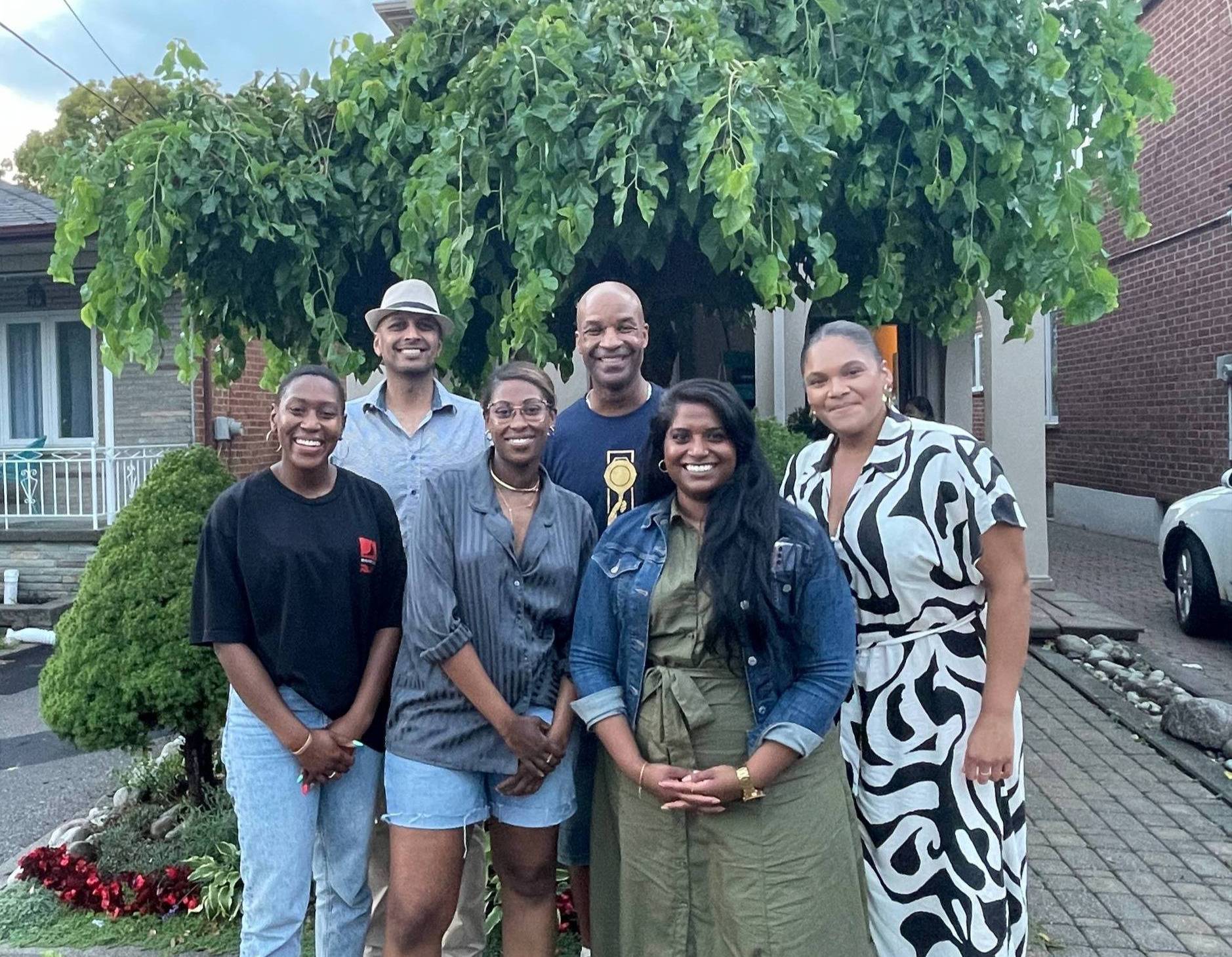 A racially diverse group of adults standing outside in front of a large tree in the summertime.