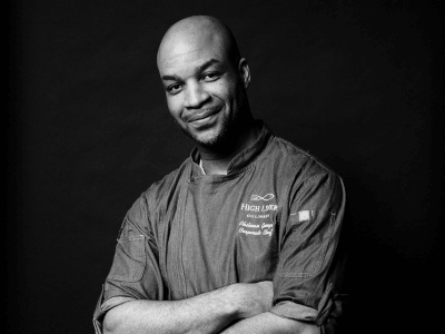 A Black man wearing a denim chef's jacket against a black background
