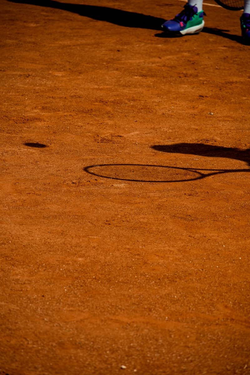 A tennis racket shadow and a ball shadow on a red clay court