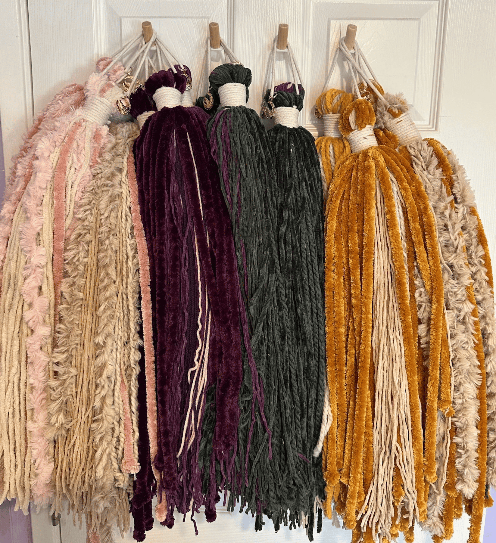 Bundles of colorful yarn, including shades of pink, beige, maroon, green, mustard yellow, and gray, are neatly hanging on hooks. Each bundle looks fluffy and textured, displayed against a white door.