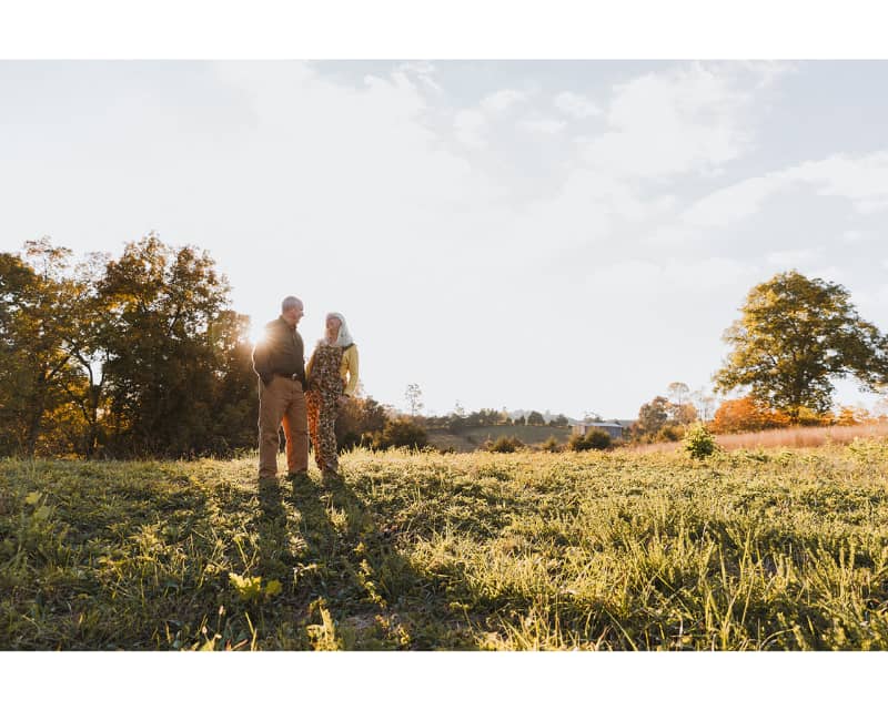 A couple standing in the middle of a fall field with the sun behind them. she is in green floral gardening overalls and He is in work pants and a green shirt.