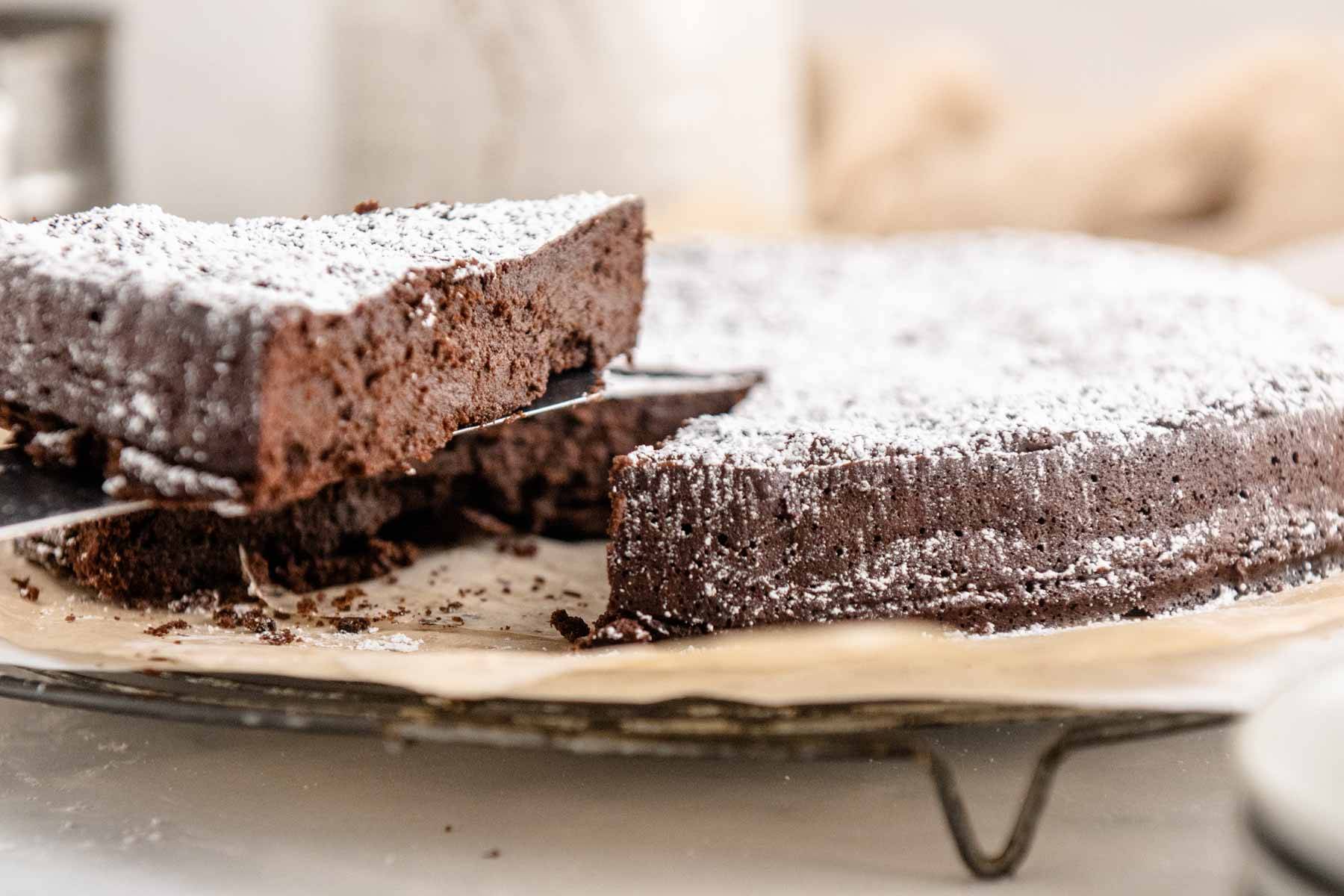 A slice of flourless chocolate cake being picked up from the rest of the torte on a cooling rack and parchment paper.