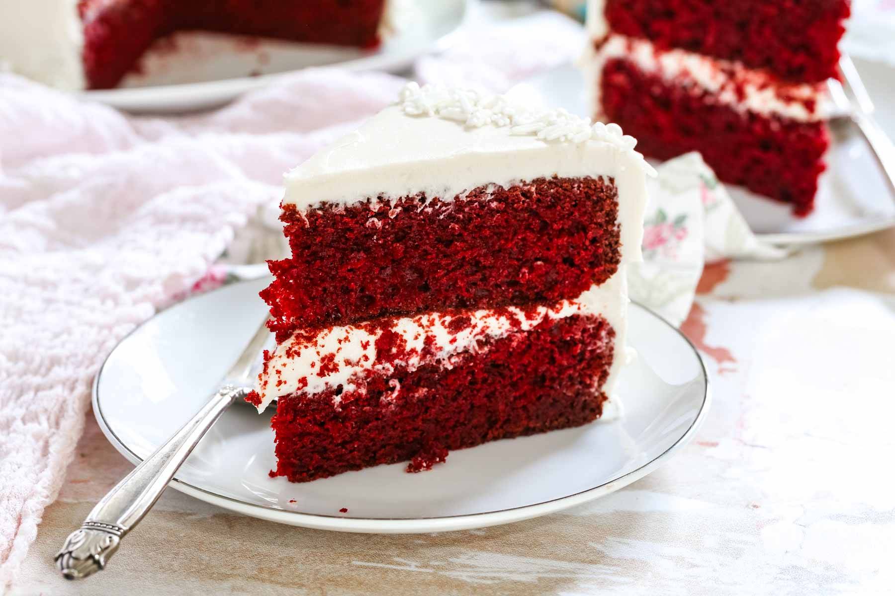 A slice of red velvet cake on a plate with another piece blurred in the background.