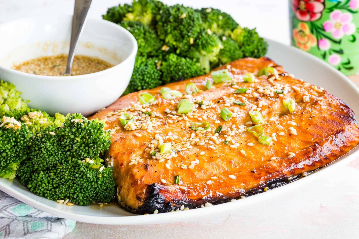 A piece of broiled salmon garnished with sesame seeds and scallions on a plate with broccoli.