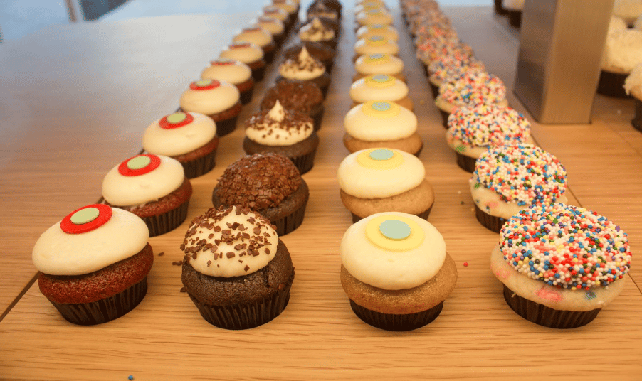 Rows of assorted cupcakes arranged on a wooden surface. The cupcakes feature a variety of toppings, including white frosting with sprinkles, chocolate pieces, and colorful circular decorations. The arrangement creates an appealing and organized display.