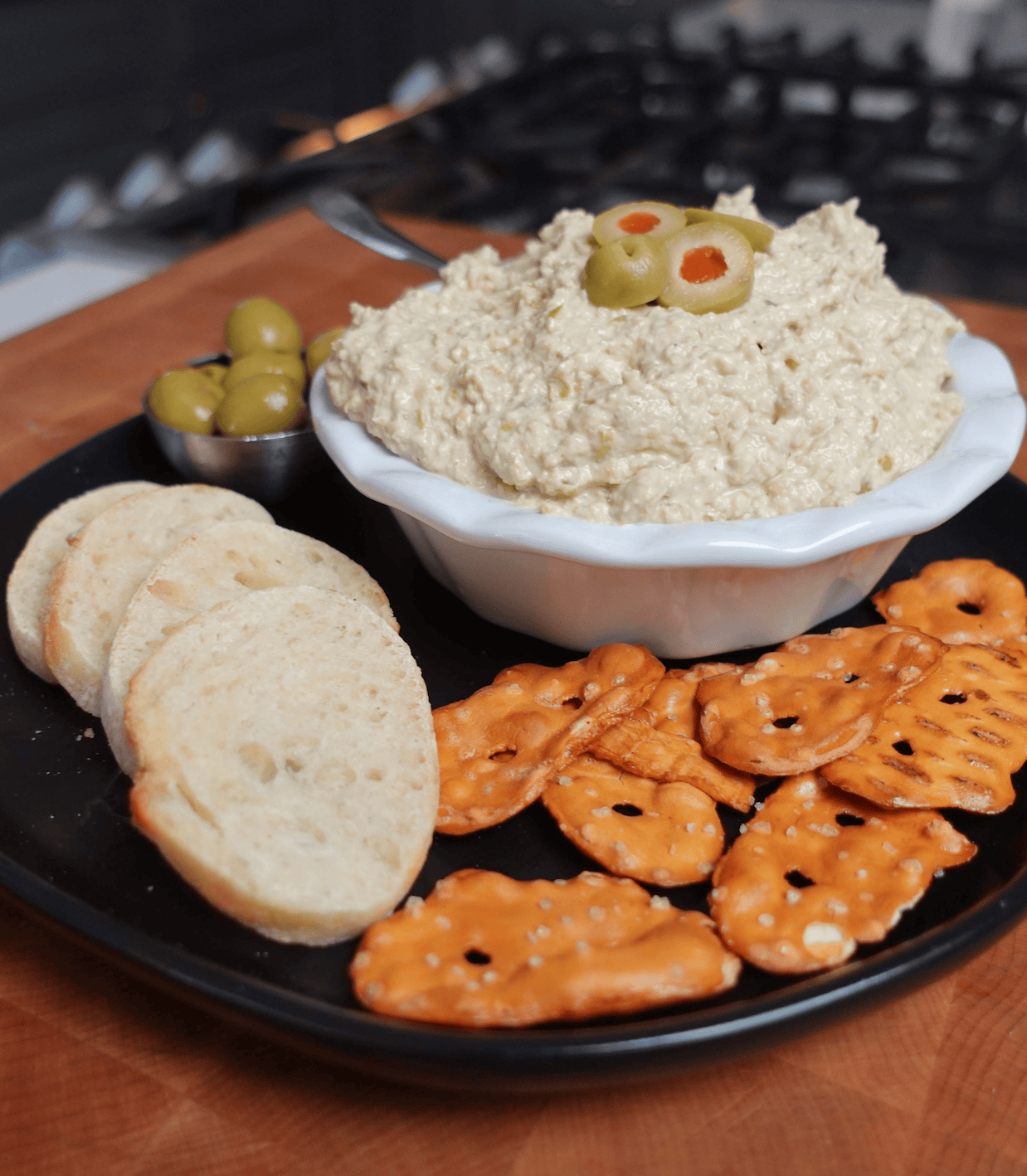 A bowl of olive dip with olives on top served with pretzel thins and crustini.