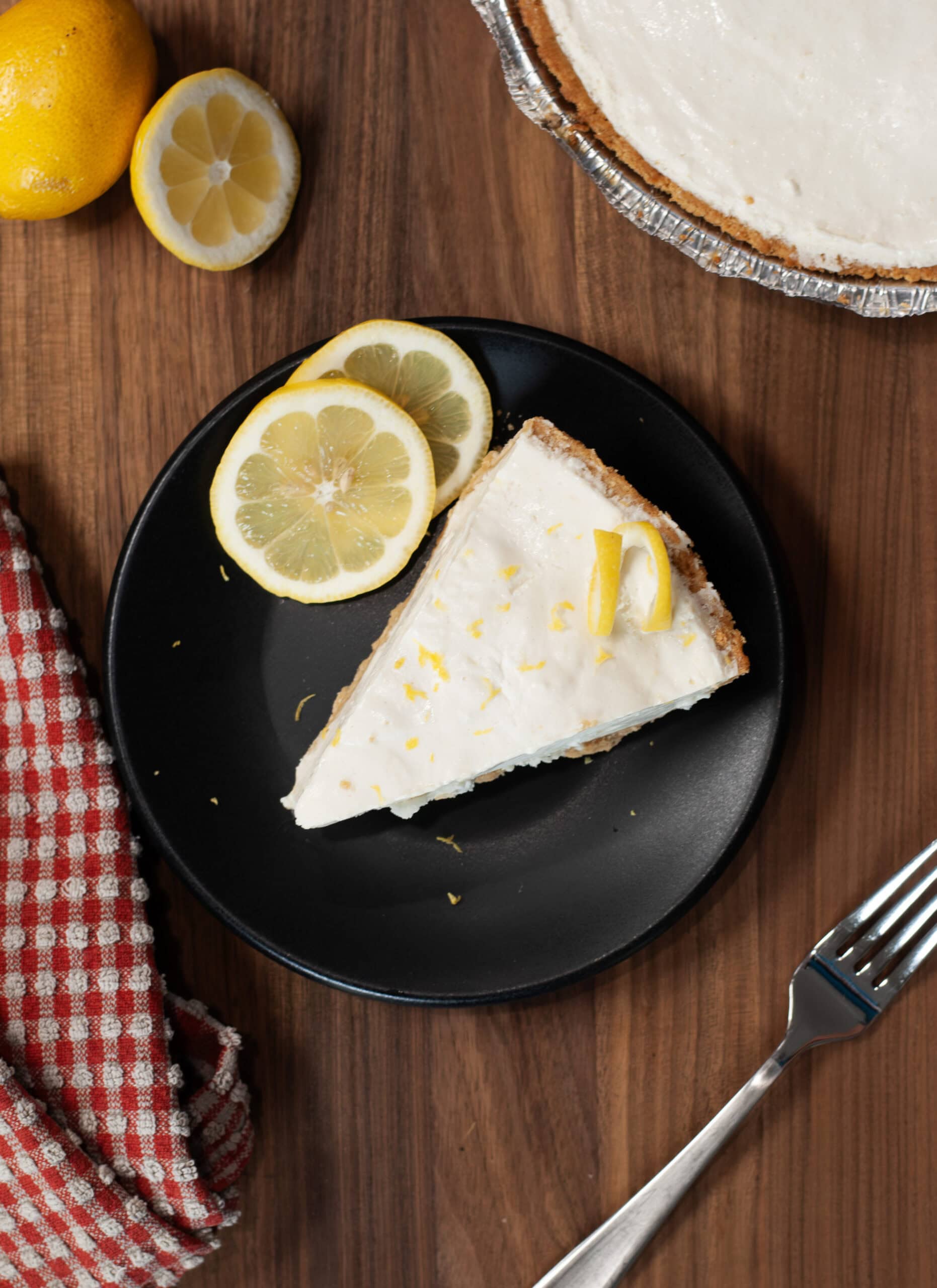 A black plate holding a slice of creamy no-bake lemon cheesecake with a lemon slice garnish. 
