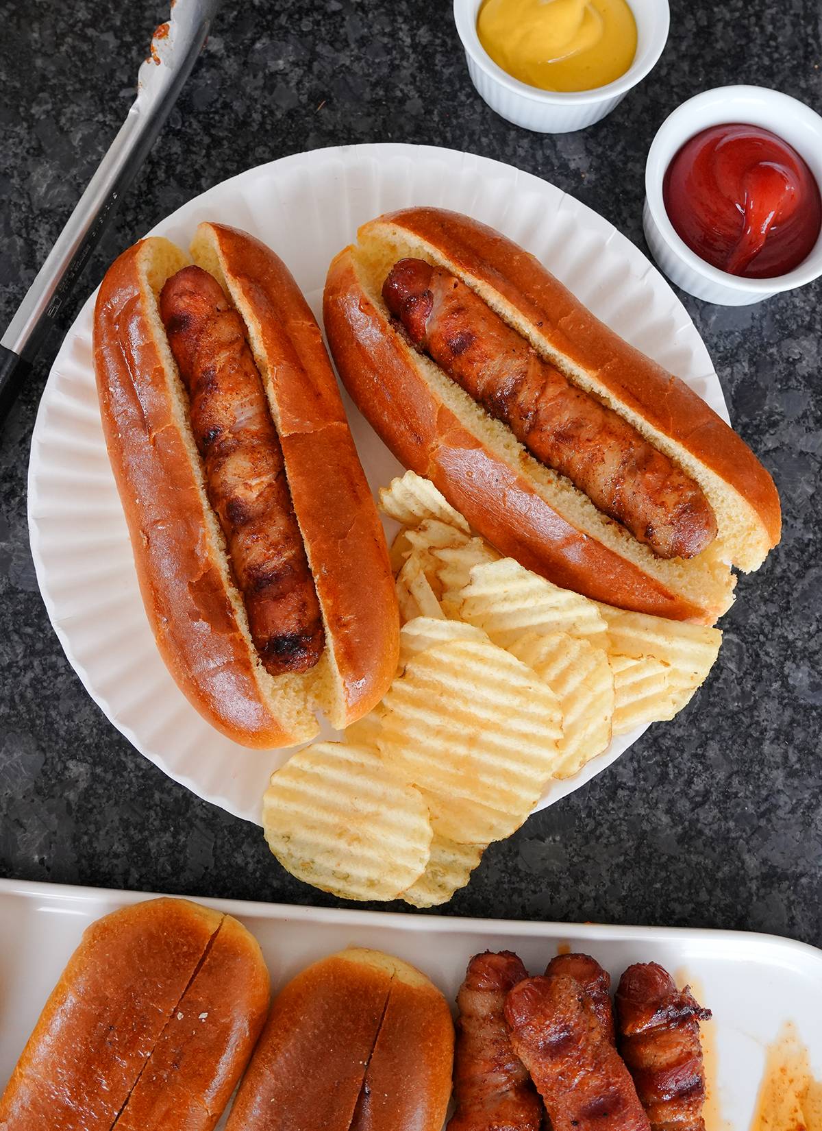 Two bacon wrapped hotdogs in buns with chips. 