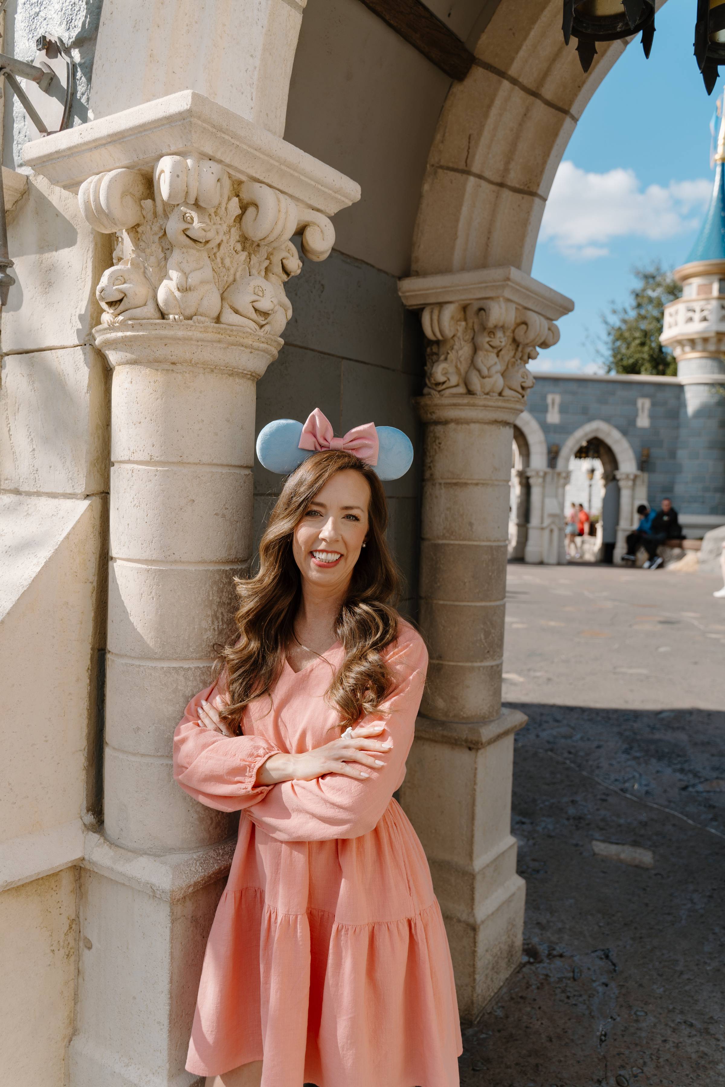 A woman wearing a pink dress and blue mouse ears with a pink bow stands smiling, leaning against a stone pillar. The setting is an outdoor scene with architectural details that evoke a castle or theme park. The sky is clear with a few clouds, and there's a blurry background with other people and structures.