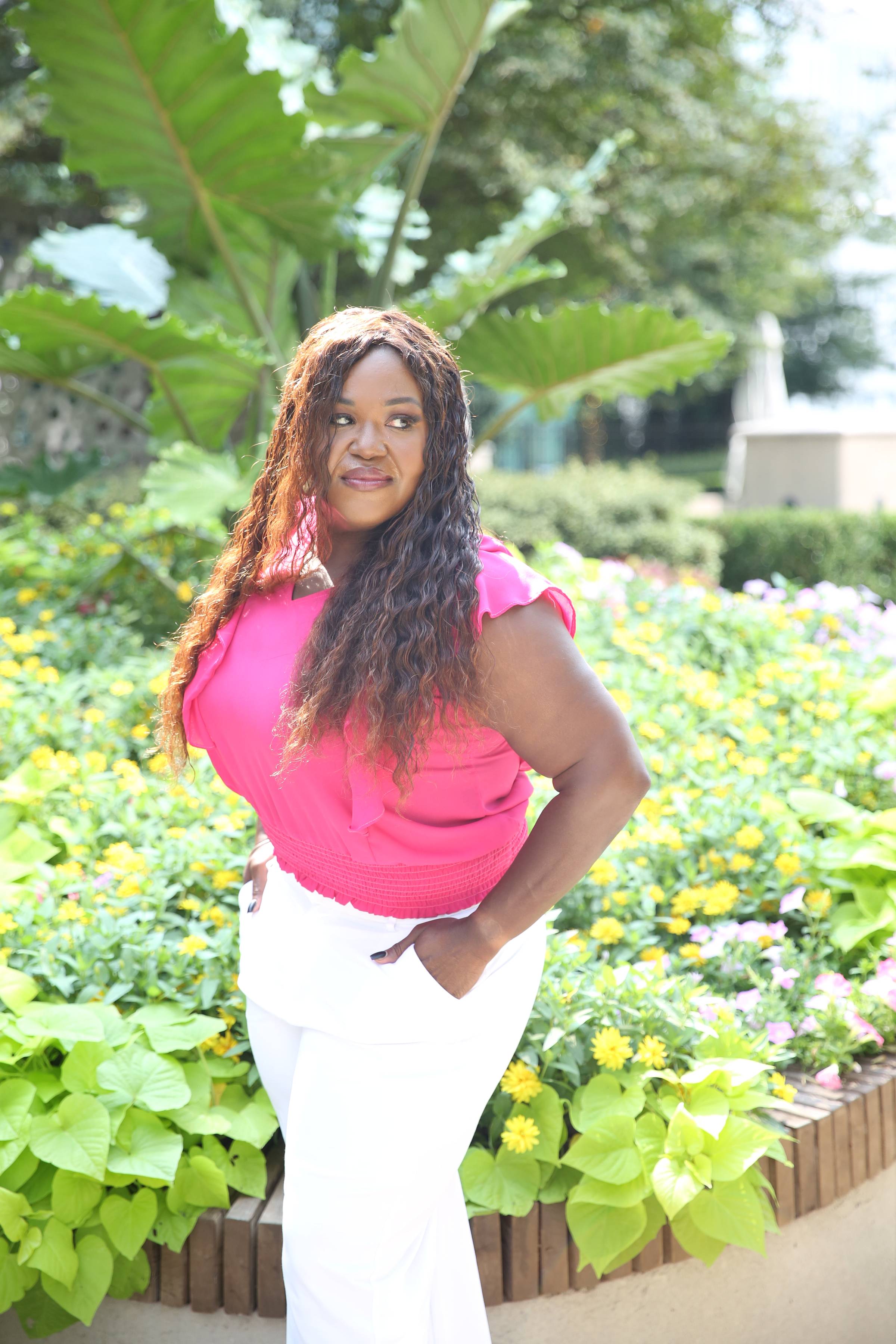 Dr. Shermnae, The Destiny Doula™, standing confidently in a vibrant pink blouse and white pants, surrounded by lush greenery and blooming yellow flowers.