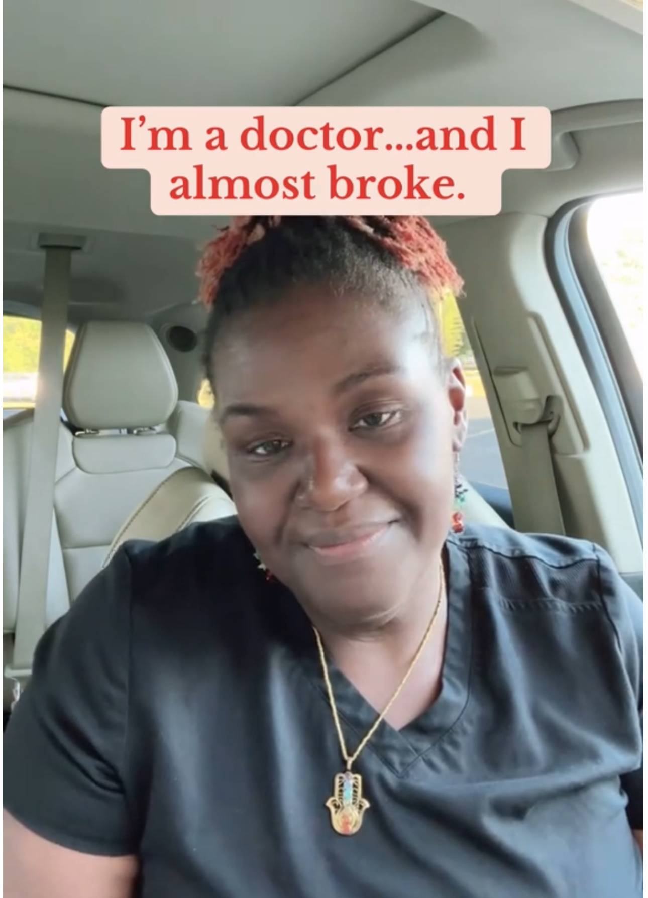 A Black woman sitting in a car wearing black scrubs, looking into the camera with a slight, reflective smile. She has natural hair pulled back in a ponytail, no makeup, and is wearing a necklace. Overlaid text on the video says, “I’m a doctor… and I almos