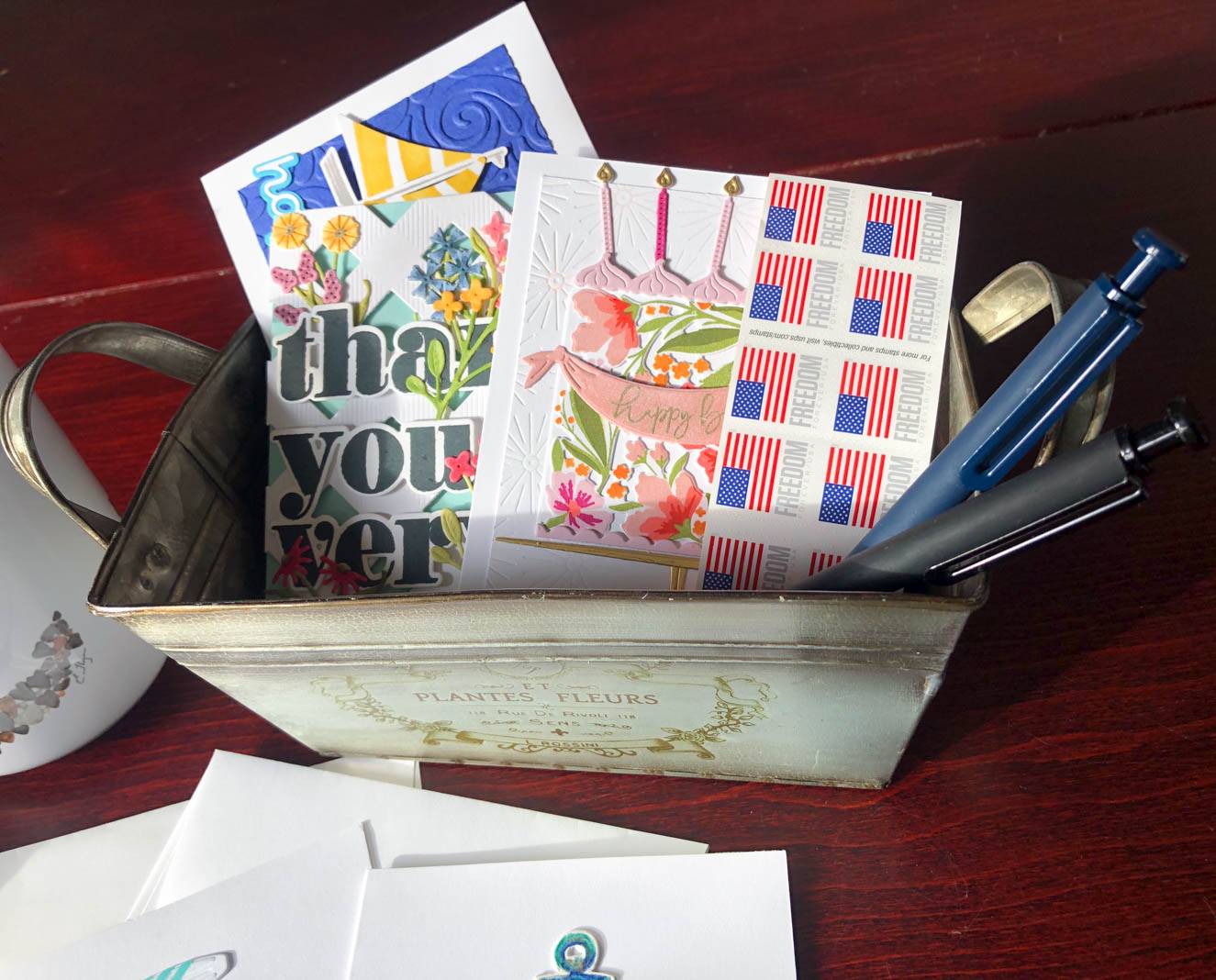 A metal container labeled "Plantes Fleurs" holds various decorative cards and a sheet of "Freedom" stamps with American flags. Inside the container are two pens. The designs on the cards include flowers, a "thank you" message, and a "happy birthday" message with candles. Envelopes and a mug are visible next to the container on a wooden surface.