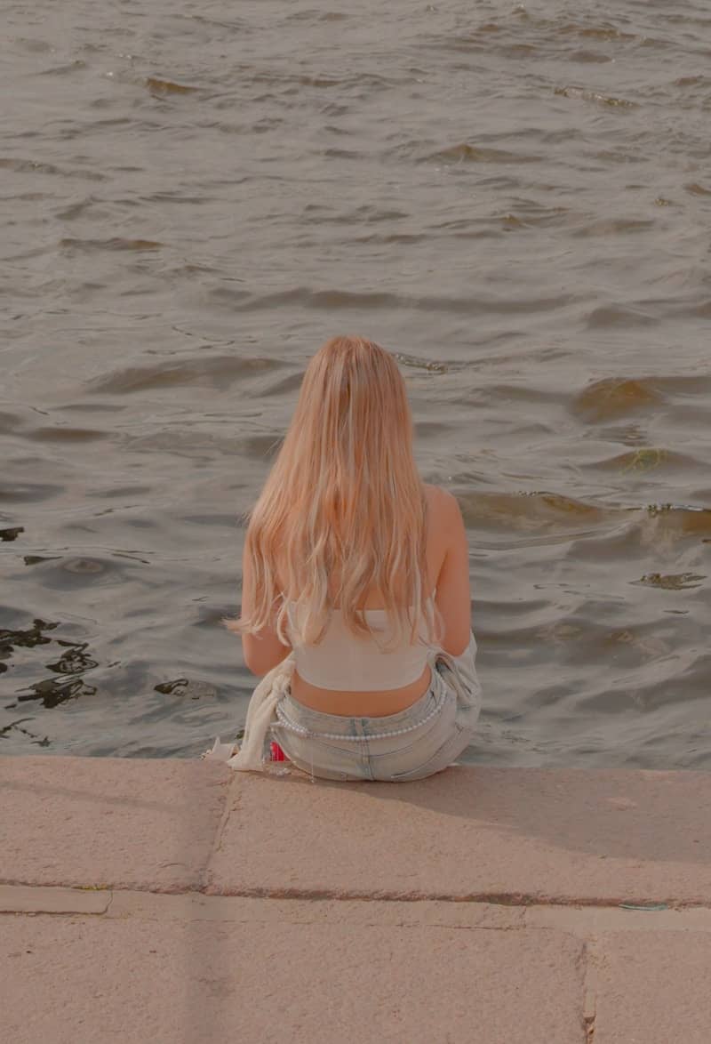 Woman with long hair sits by the water.
