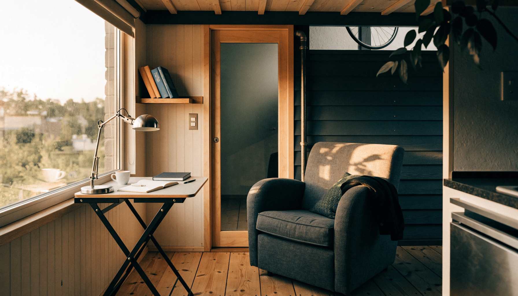 A minimalist tiny home living area featuring a fold-down desk, a wall-mounted shelf with a few carefully chosen books, and a cozy armchair bathed in soft afternoon light.
