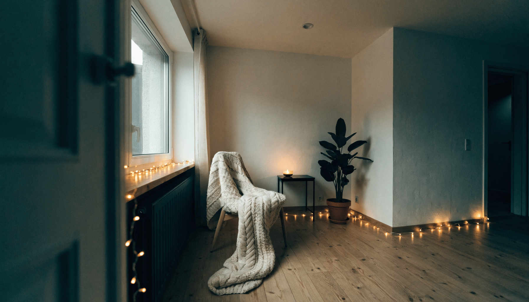 A snug minimalist corner with a chunky knit blanket draped over a chair, a small lit candle, a potted plant, and warm string lights glowing softly.