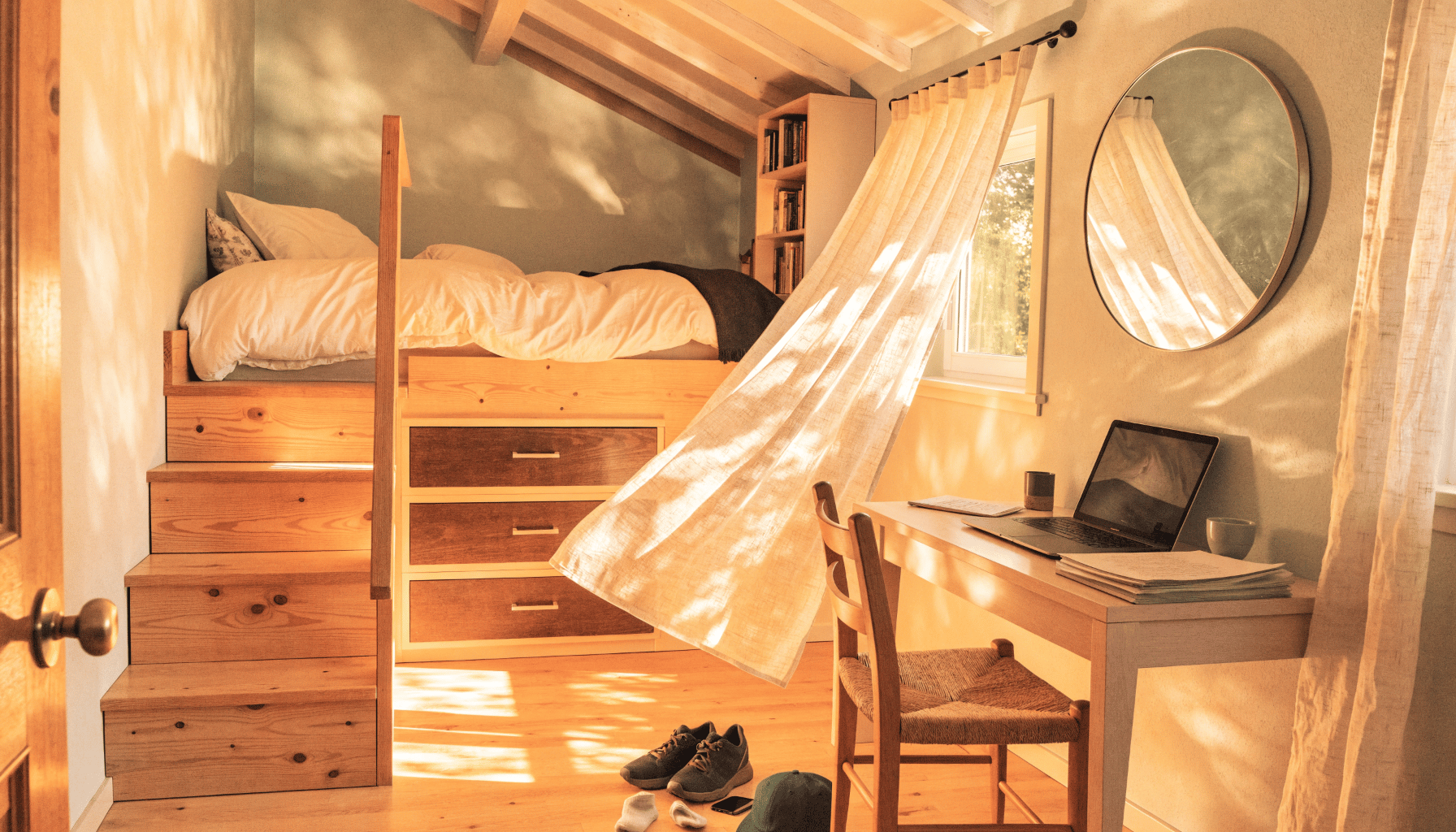 A bright tiny bedroom with a lofted bed, built-in drawers beneath the stairs, a large round mirror, and flowing sheer curtains.