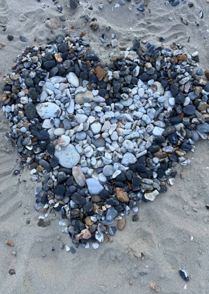 A heart shape formed by smooth grey and white pebbles on sandy beach.