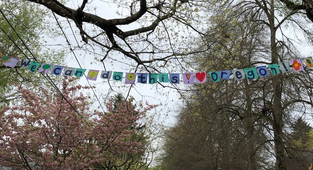 A photo of a banner made of individual square pieces of cloth that spell out we can to this Oregon. You see it against a backdrop of tree branches.