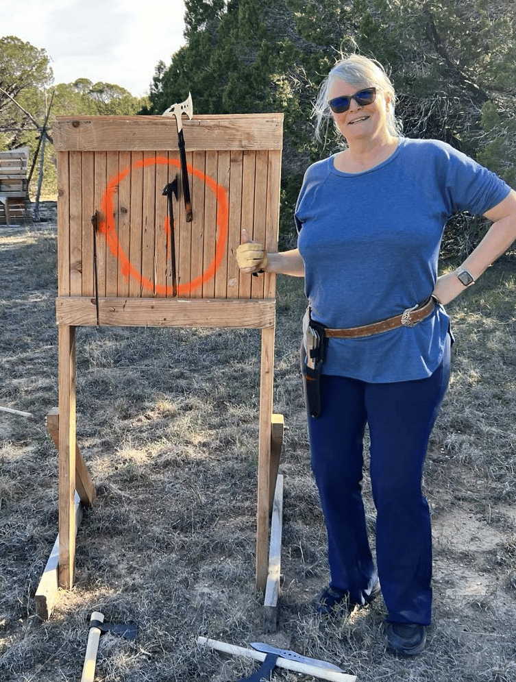 Stocky older woman with gray hair tied back, but blowing in the wind, smiling with darkened glasses, wearing a blue shirt and pants with a leather belt with a knife sheath hanging from it standing next to a wooden target with an orange circle painted on it and three axes stuck at random places on the target.