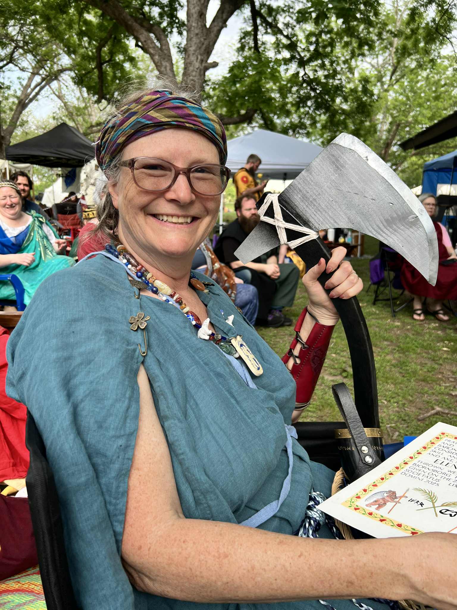 Paige with big grin wearing a light blue roman tunica, a stripey colored head cloth, and a red bracer while holding a huge axe with a hand-painted award scroll in her lap. Other folks in medieval garb with shade canopies are in the background.