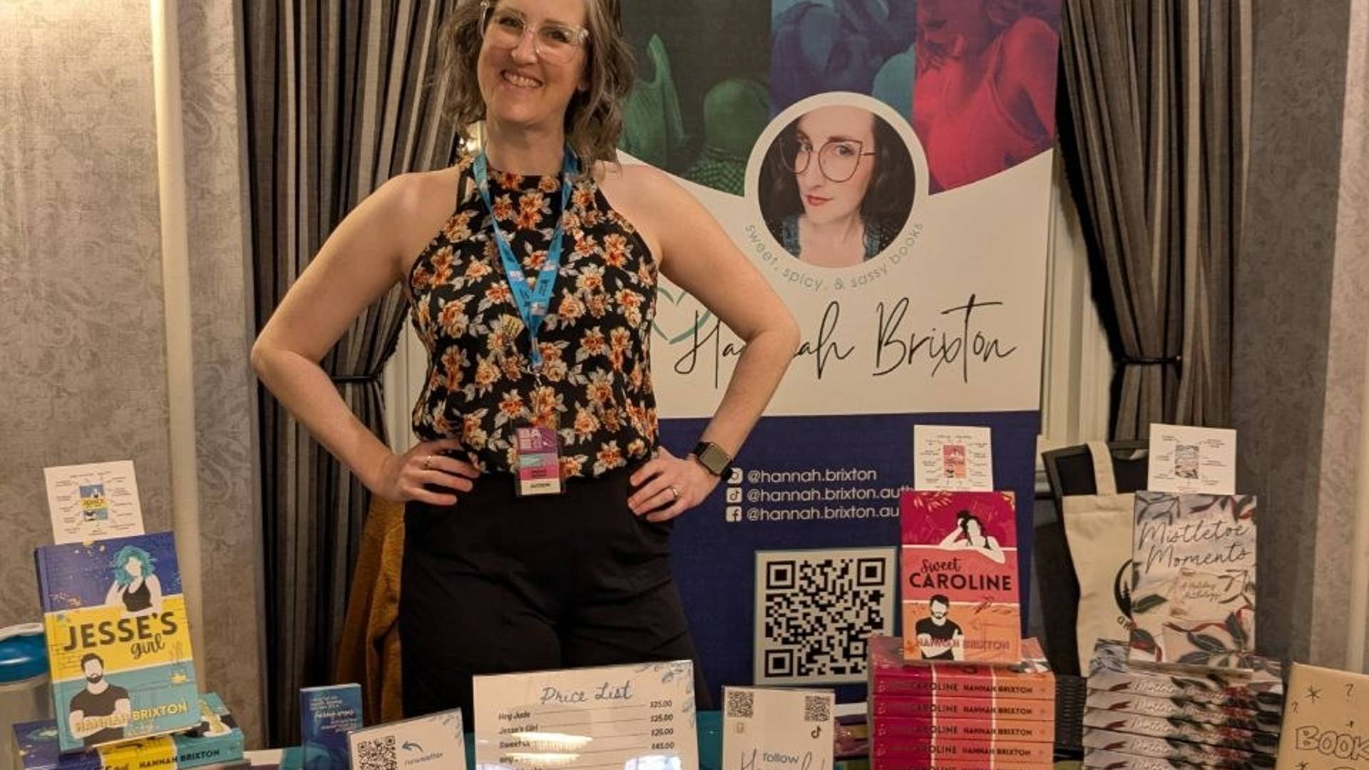 Romance author Hannah Brixton smiling behind her author table at Bonded and Enchanted, surrounded by stacks of her books and a banner with her name on it