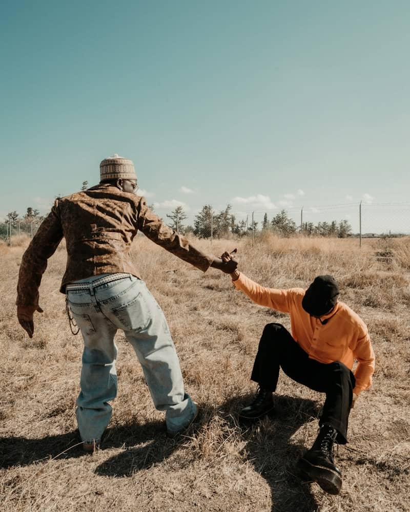A person in a patterned jacket helping another person in an orange shirt up from dry grass
