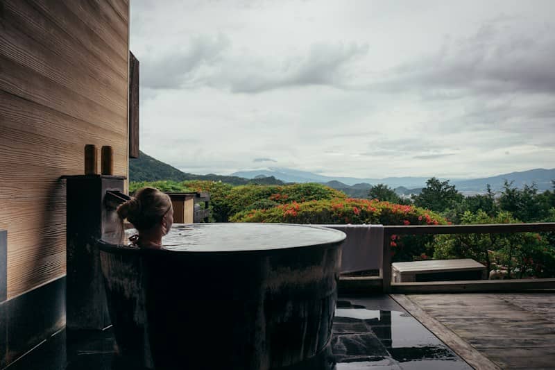 Woman relaxing in an outdoor wooden bathtub with mountain view.