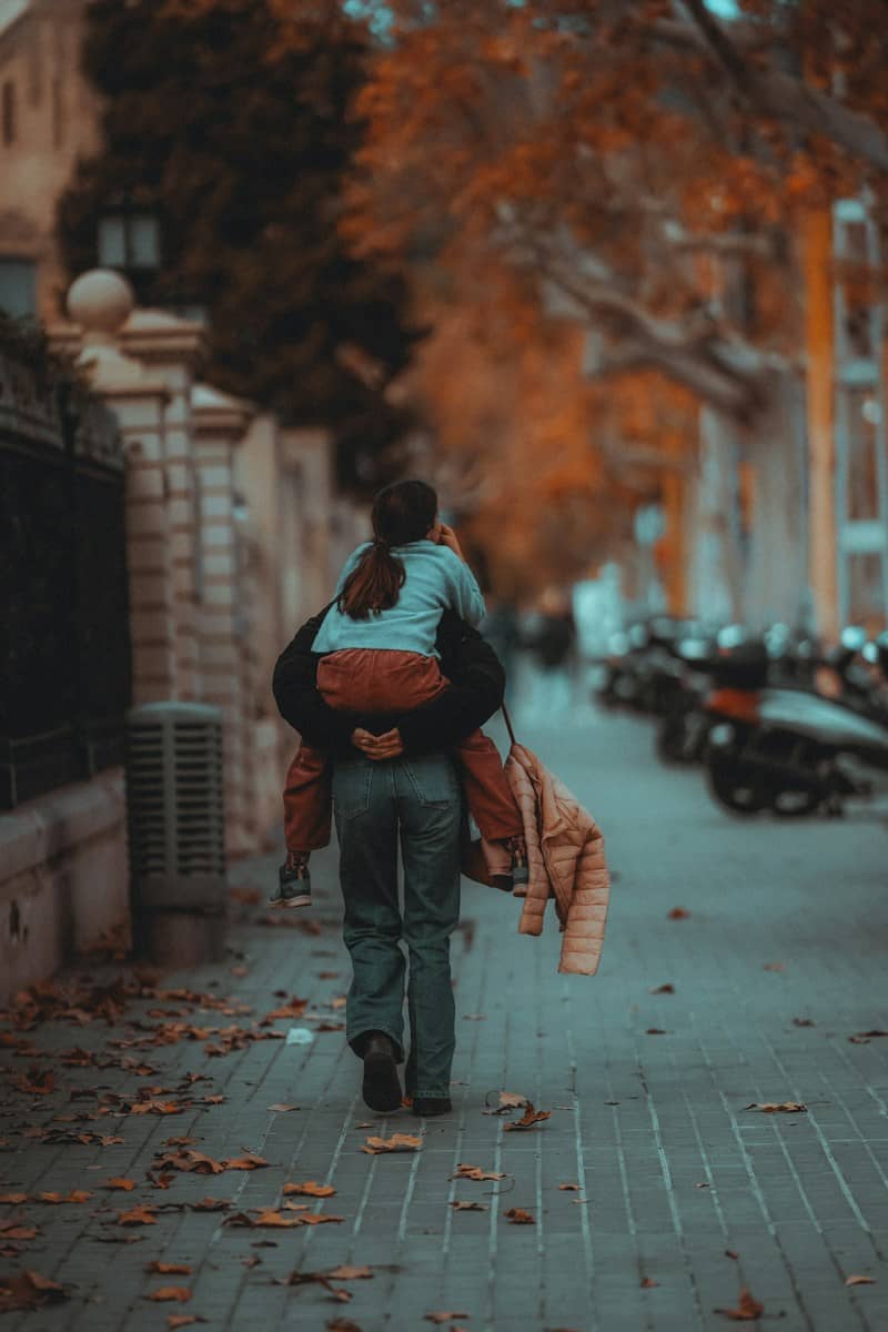 An adult carrying a child on their back along a tree-lined sidewalk in autumn