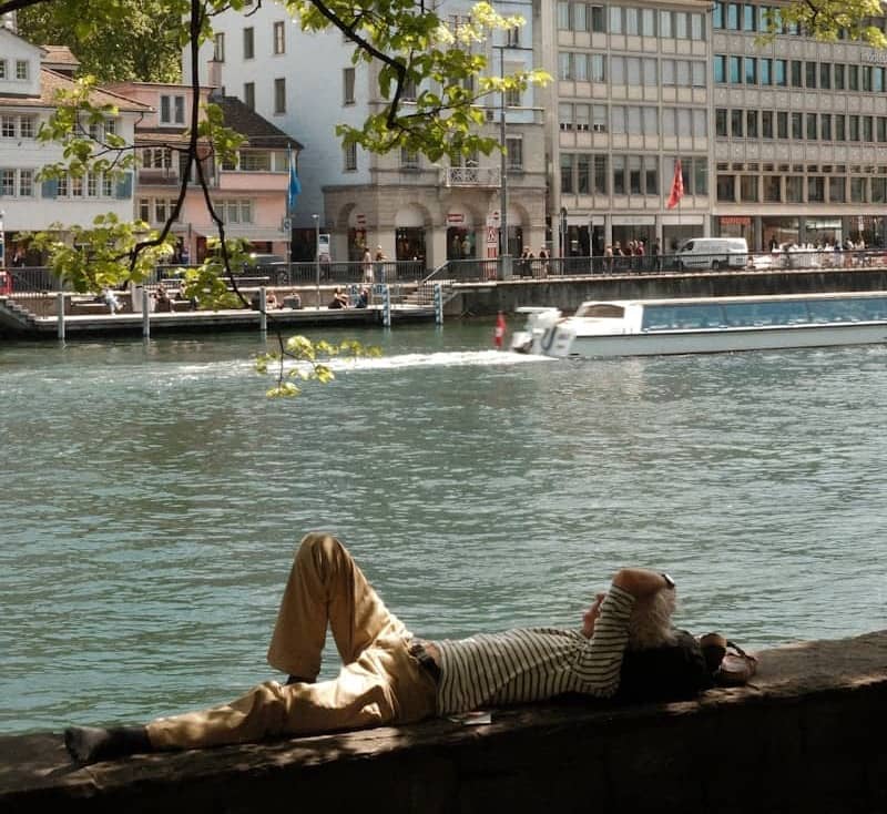 Person lying by river, city buildings and boat in background.
