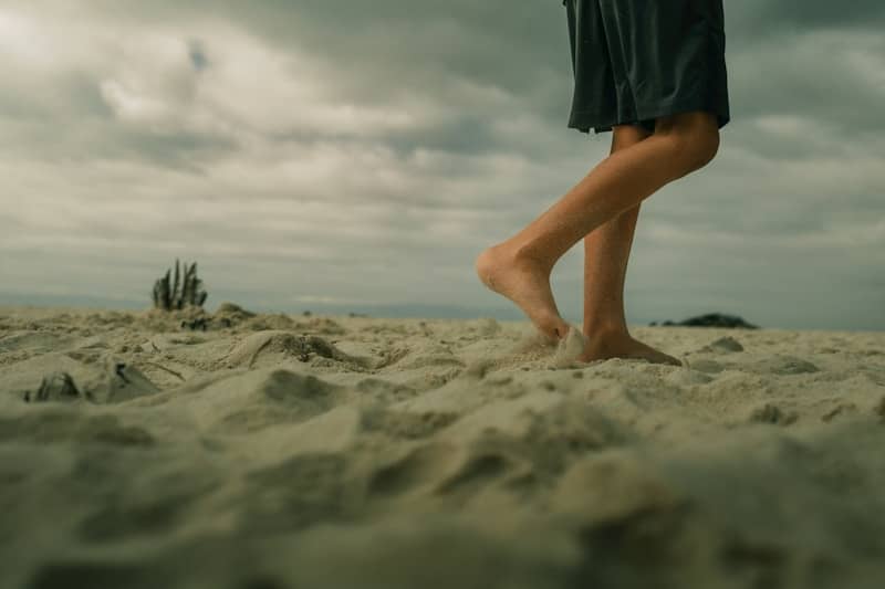 Barefoot person walks on sandy beach under cloudy sky.