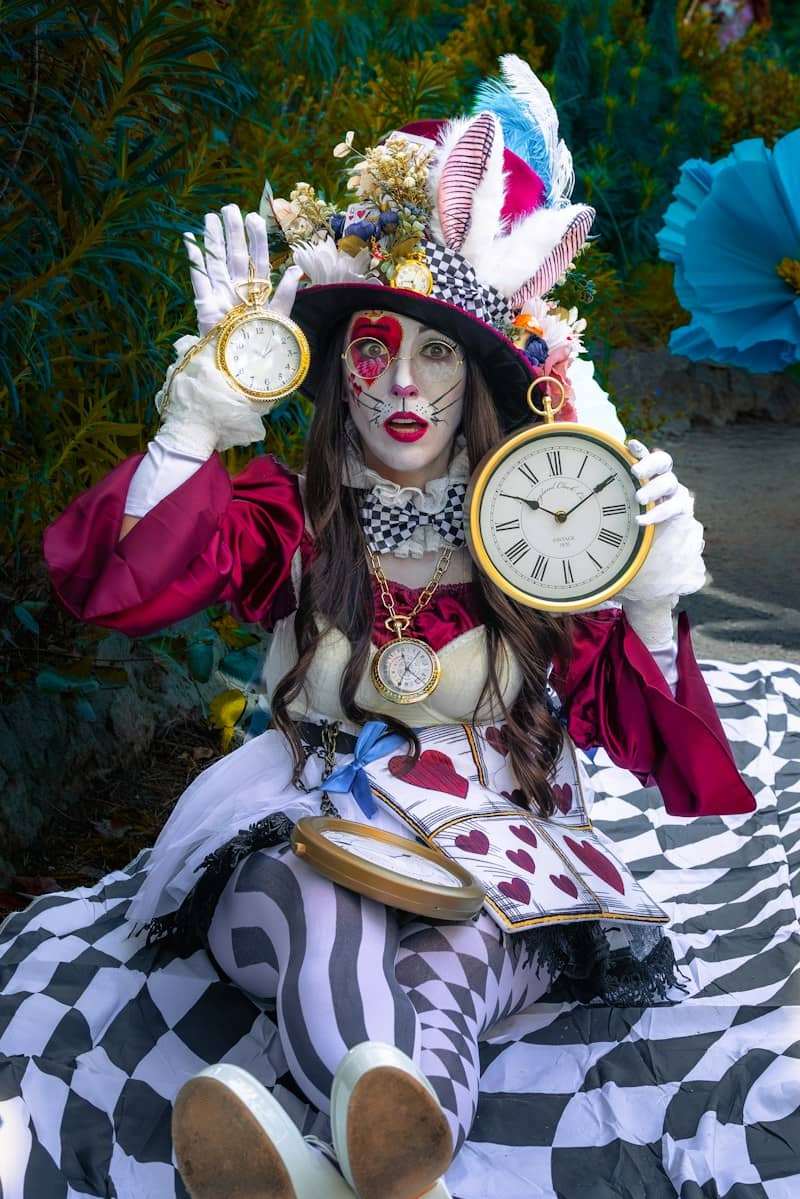 A person in a Mad Hatter costume holding two clocks while sitting on a checkered blanket