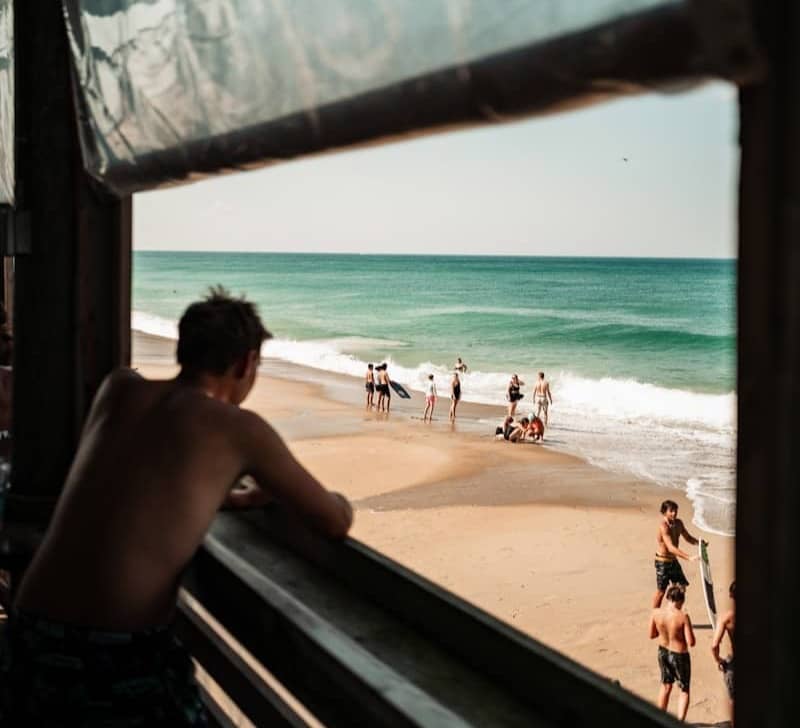 Man watches people on a sunny beach from a pier.