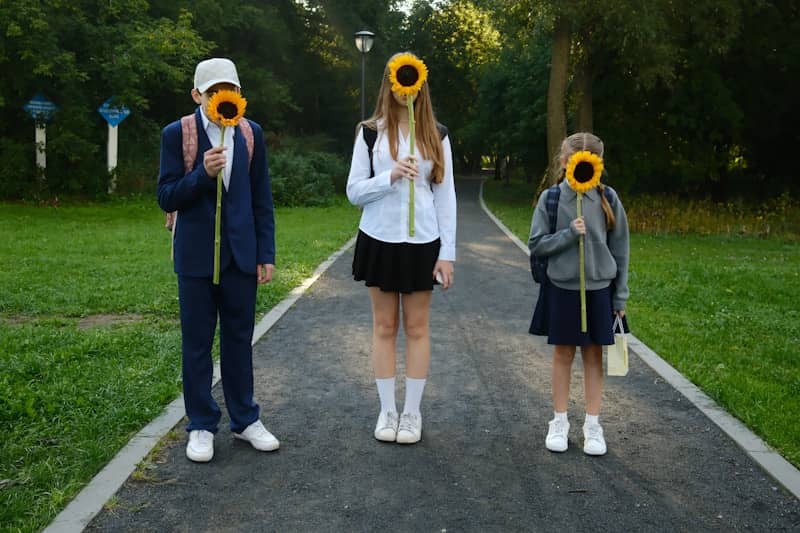 Three students holding sunflowers in front of their faces on a park path