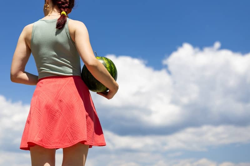Woman in red skirt holding a watermelon under blue sky.