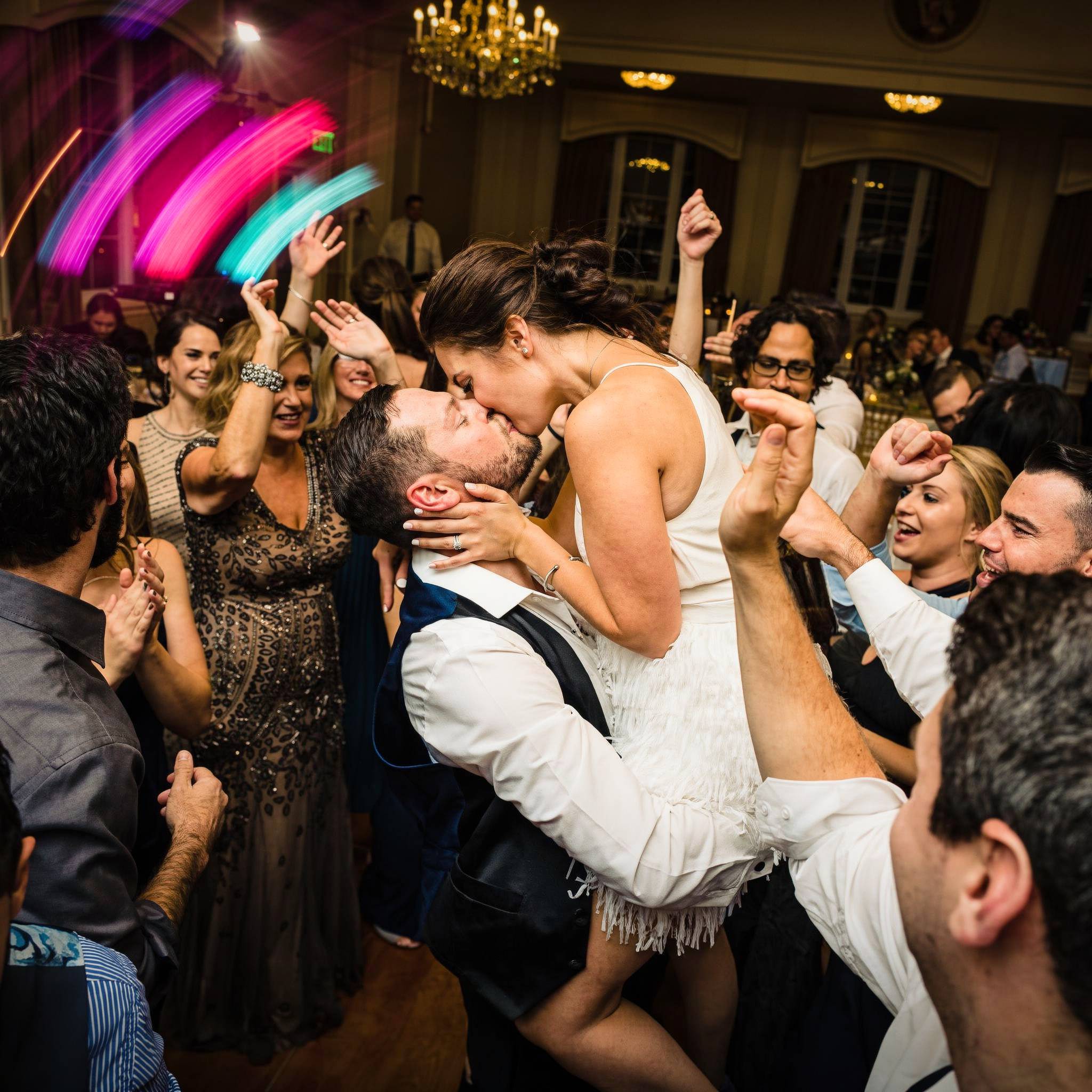 Wedding guests toast the kissing couple with raised drinks.