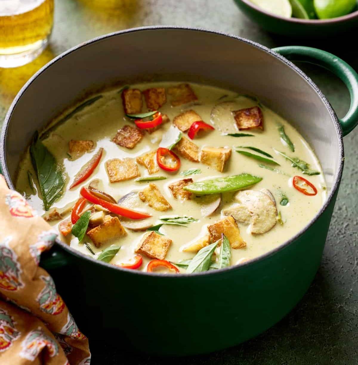 backlit shot of Thai vegan green curry with crispy tofu and vegetables in a green saucepan with beer and limes in background.