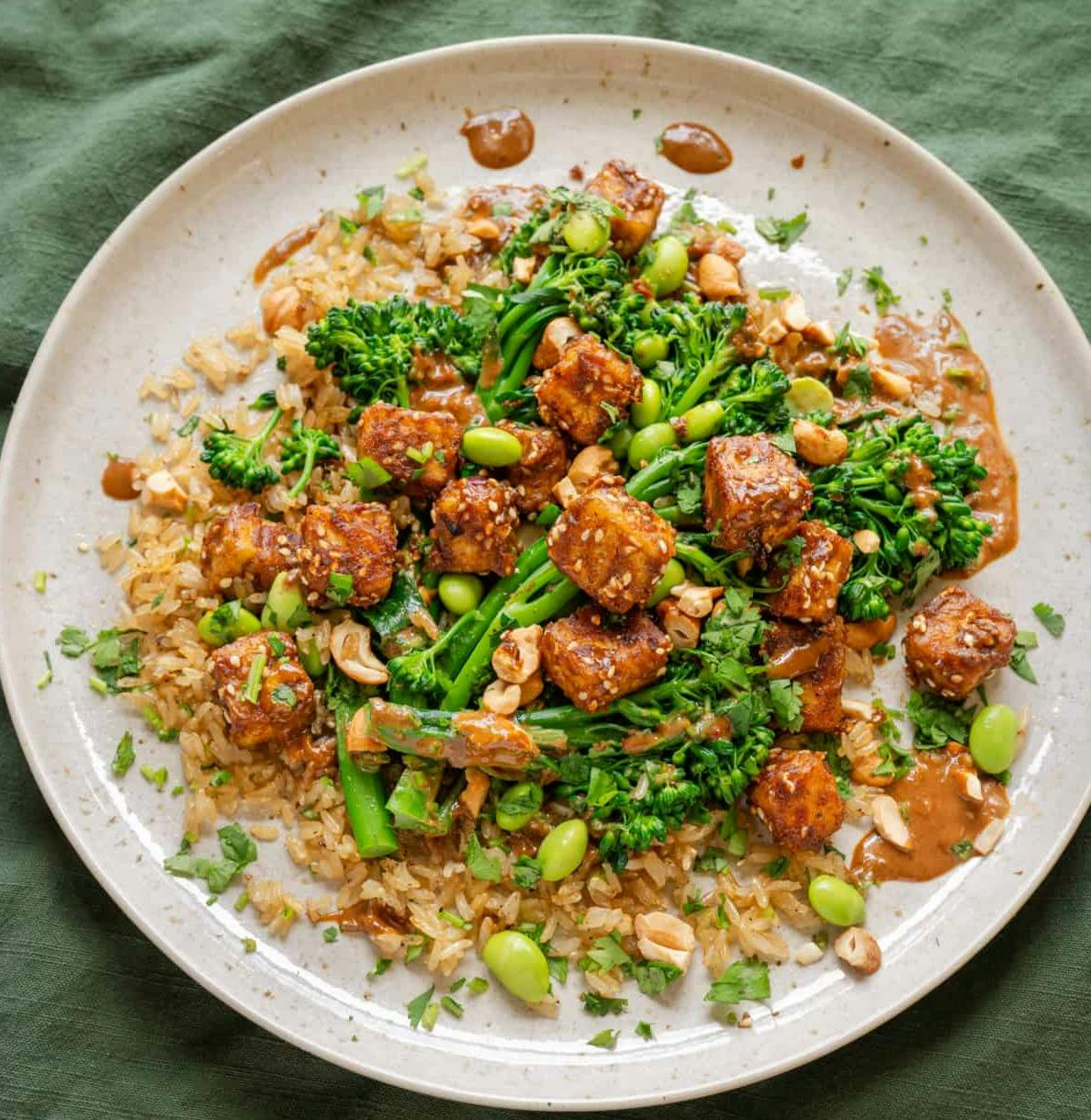 baked peanut tofu with broccolini and edamame on top of brown rice on a ceramic plate.