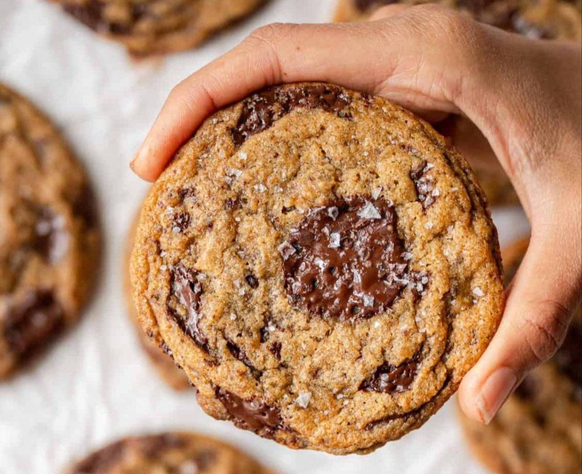 woman's hand holding large vegan chocolate chip cookie with other cookies in background.