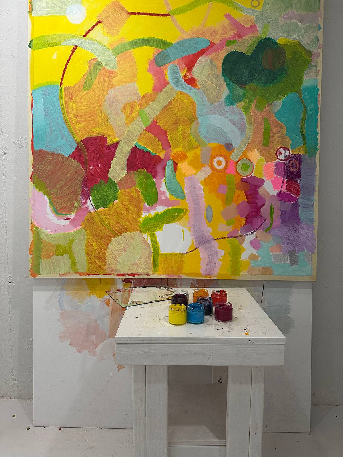 An abstract painting sitting on a makeshift easel. There's a table with jars of various paint colors in front of the painting