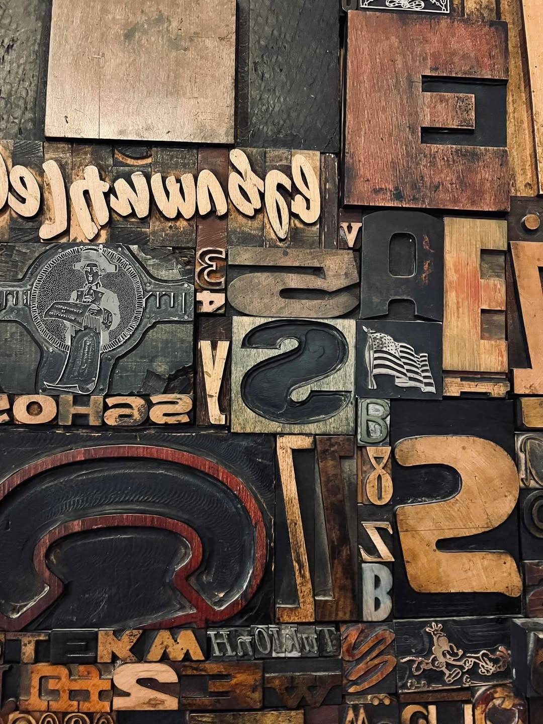 A collection of vintage wooden printing blocks arranged closely together. The blocks display a variety of letters, numbers, and symbols in different fonts and sizes. Some blocks include decorative elements and intricate engravings. The assortment showcase