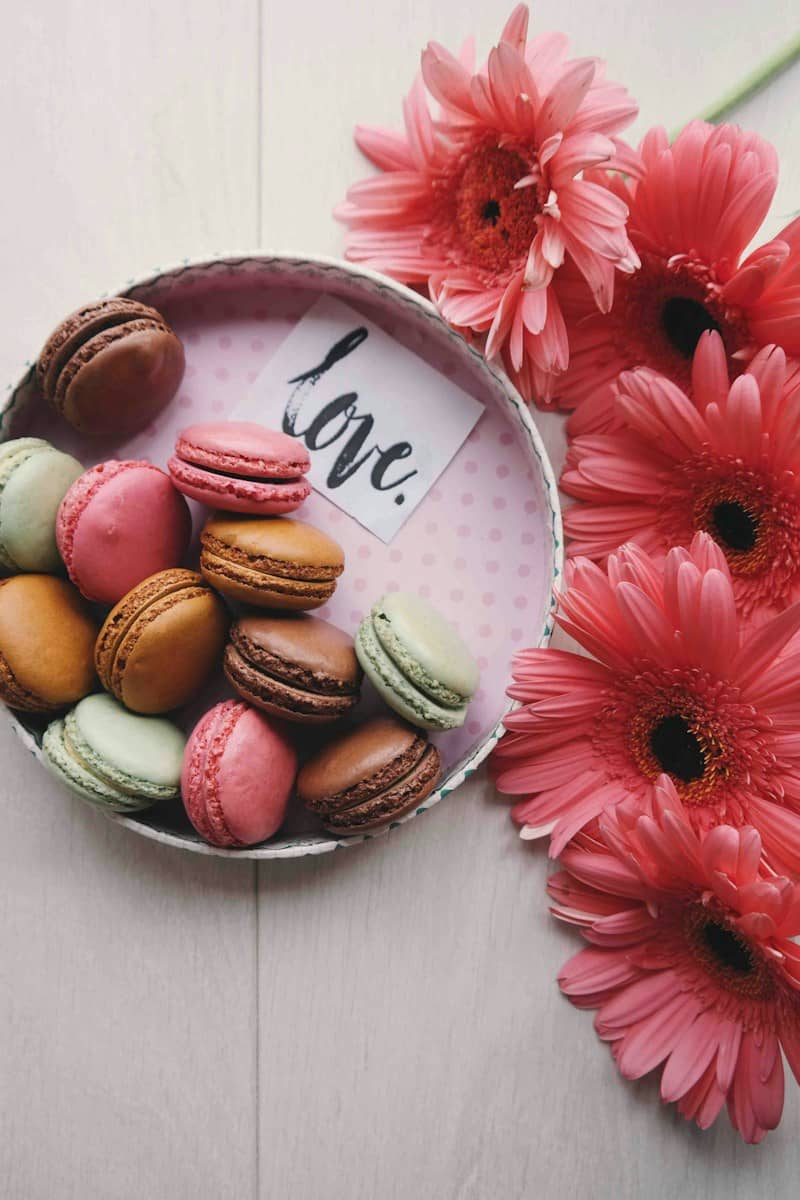 Pink gerber daiseys laying next to a small white wicker bowl that holds brown, yellow, pink, and green macarons and a note with the word "love"