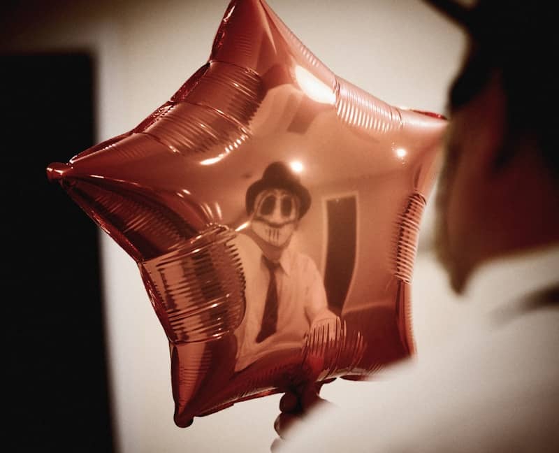 An out of focus person holding a star-shaped balloon. In the balloon, you can see the reflection of  a painted face wearing a white shirt, black tie, and hat. Please never ask how long I looked for a picture that was spooky but not going to give my reader