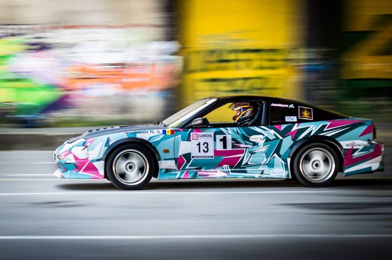 A super colourful racecar with hot pink, light and dark blue, and white geometric patterns against a blurry background (because the car is going so fast, presumably). The car has the number 13 on the door