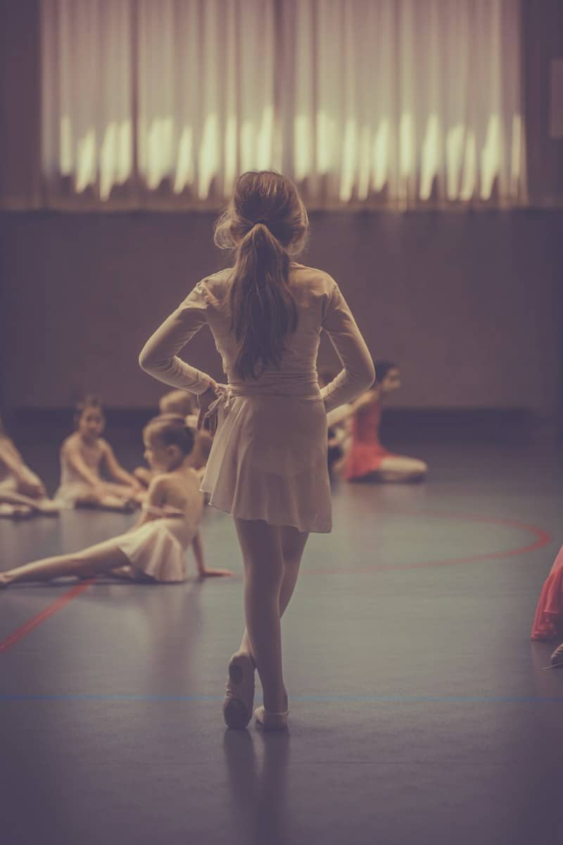 A girl with a ponytail standing with her feet crossed in a ballet costume. Other children in ballet costumes are on the floor around her..