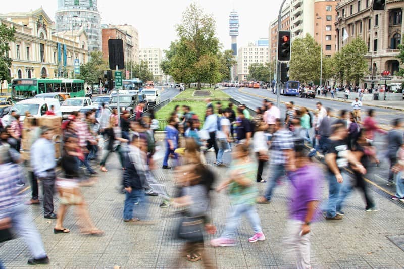 Blurred, colourfully-dressed people walking on a sidewalk between buildings with cars passing in the background.