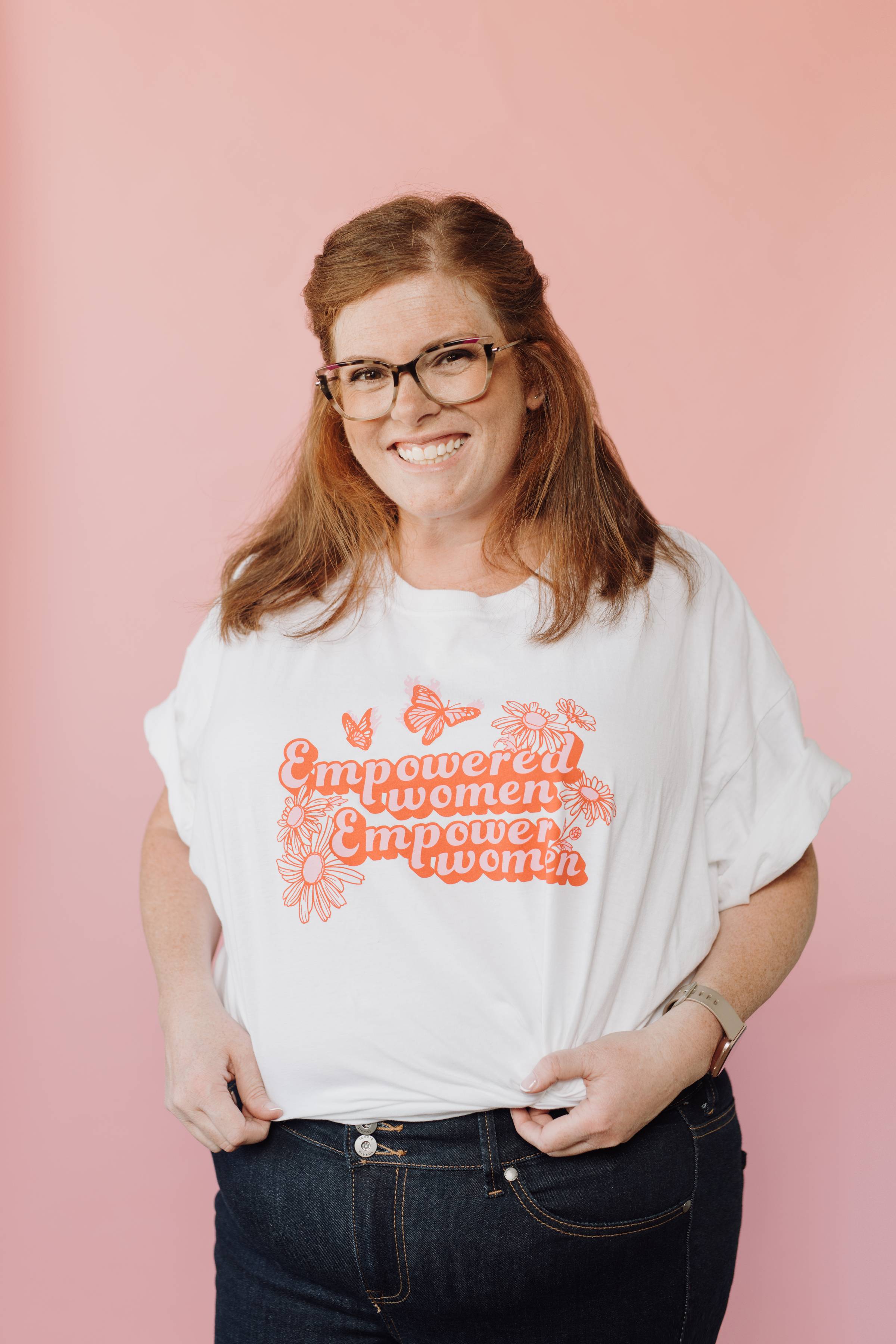 Allison, a redhead with glasses, has a big smile wearing a white shirt that says "Empowered women empower women" in two-toned pink lettering, with flowers and butterflies around the words. She's standing in front of a pink background.