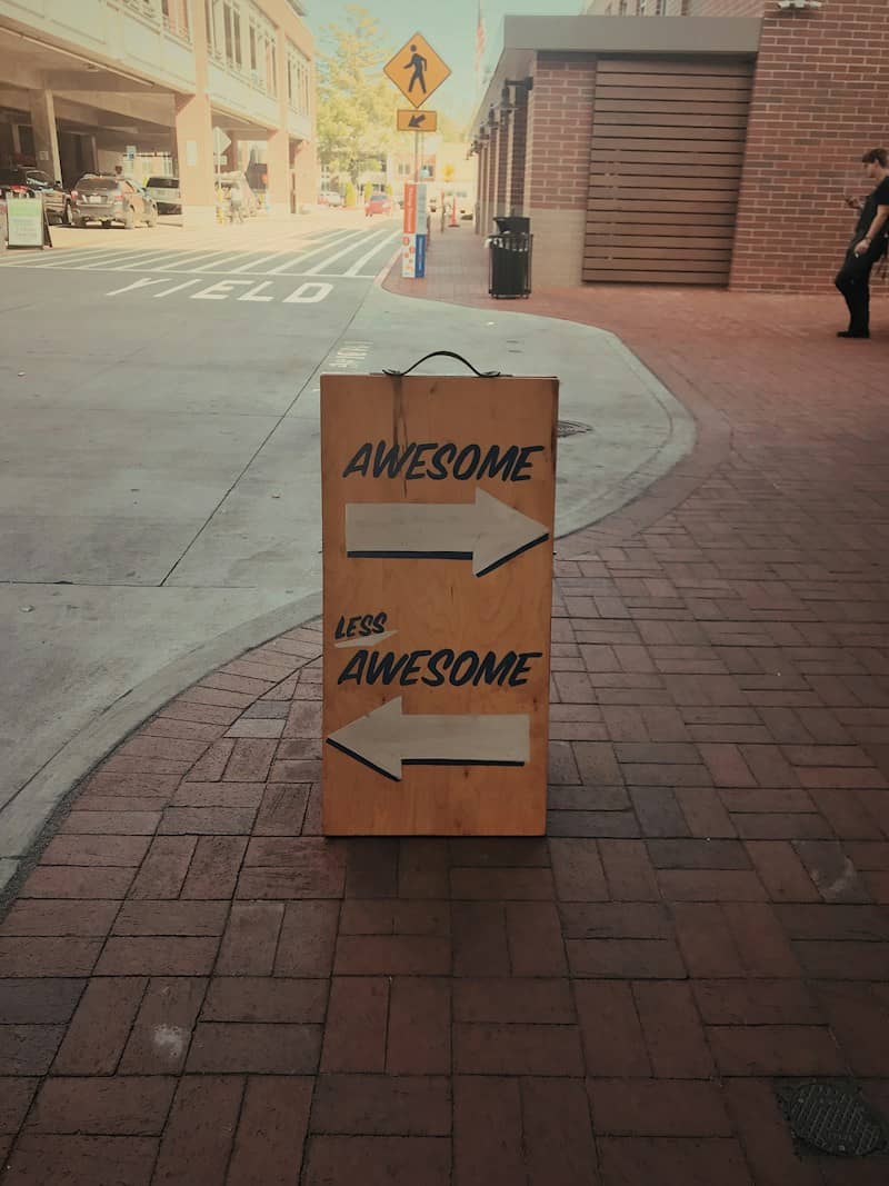 A wooden sandwich board on a brick sidewalks. The word "awesome" with an arrow pointing to a right, and "less awesome" with an arrow pointing to the left. There's a person looking at their phone in the distance and a yield sign on the street beyond the sign.