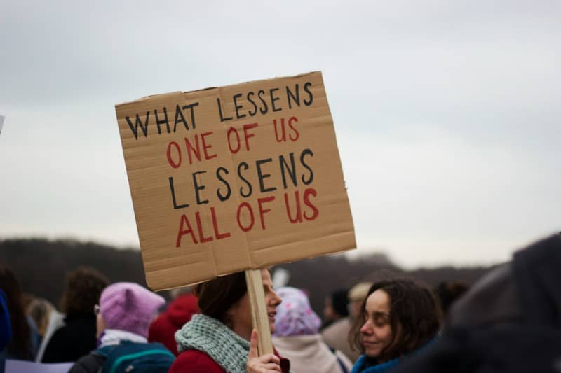 Heads and shoulders of women at a demonstration. One is holding a cardboard sign that says "what lessens one of us lessens all of us".