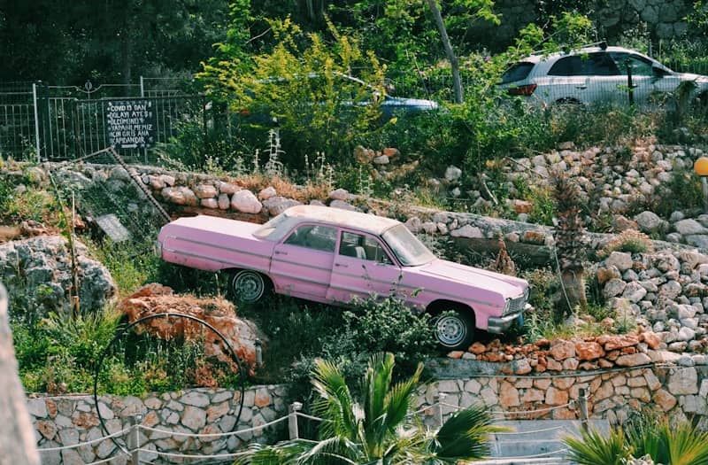 A pink classic car sitting among grass and rocks on the side of a hill, with no street in sight.